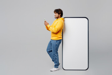Full body side view young Indian man wear casual yellow hoody stand near big huge blank screen mobile cell phone with workspace area use smartphone isolated on plain grey background studio portrait. - Powered by Adobe