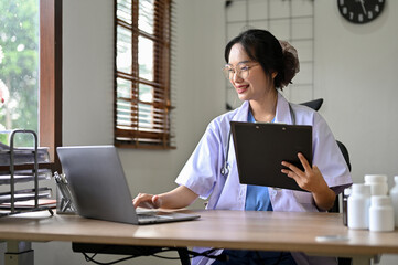 Beautiful Asian female doctor using laptop computer, working at her desk in the doctor's office.