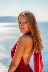 Naklejka premium Smiling young woman in a red dress looks at the camera. A beautiful tanned girl enjoys her summer holidays at the sea. Portrait of a stylish carefree woman laughing at the ocean.