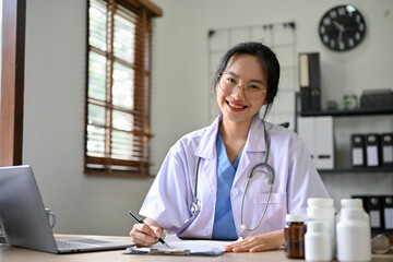 Beautiful Asian female doctor in uniform sits at her desk, smiling and looking at the camera.