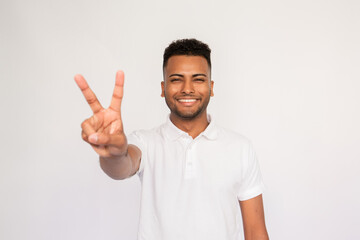 Naklejka premium Portrait of happy young man making victory gesture over white background. Latin American guy wearing white T-shirt looking at camera and smiling. Success and happiness concept