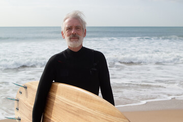 Portrait of serious aged man with surfboard. Thoughtful bearded man standing on sea beach holding...