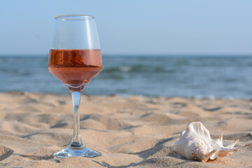 Glass of tasty rose wine and seashell on sand near sea, space for text
