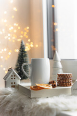 Eine Tasse Tee und Kekse auf einem weiße Lammfell an einem Fenster. Lichterkette, Weihnachten.