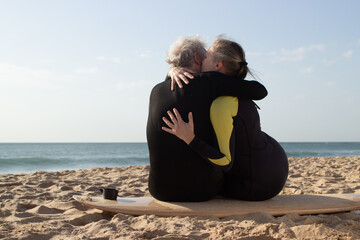 Back view of happy aged couple kissing on surfboard. Grey-haired man and woman in wetsuits sitting close on wooden board on sea beach and gently hugging. Relations, active rest of aged people concept