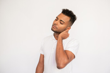 Overworked young man feeling pain in his neck. Male Indian model with closed eyes and curly hair in casual clothes touching his neck feeling discomfort, sitting too much. Physical pain concept