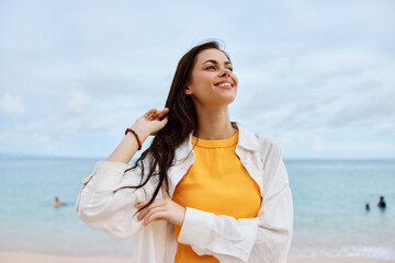 Happy woman smile with teeth with long hair brunette walks along the beach in a yellow t-shirt denim shorts and a white shirt near the sea summer journey and feeling of freedom, balance