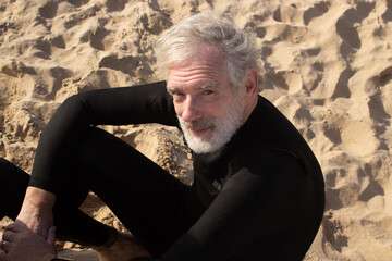 Portrait of aged male surfer resting on sand. Happy bearded man in wetsuit sitting on beach...
