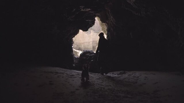 A man is walking out of a beach cave followed by his little dog