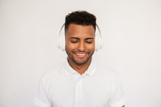 Joyful Young Man Listening To Music In Headphones Looking Down. Male Indian Model With Closed Eyes And Curly Hair In White Polo Shirt Smiling Wearing Headphones Enjoying Music.Lifestyle, Music Concept