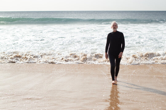 Serious aged man having active rest near sea. Sporty grey-haired man in wetsuit going out of sea after swimming walking barefoot looking at camera. Sport and healthy lifestyle of aged people concept
