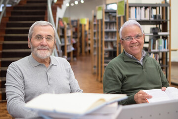 Portrait of joyful elderly men enjoying reading in library. Two smiling men sitting at table reading books about technologies getting new knowledges, looking at camera. Education when retired concept