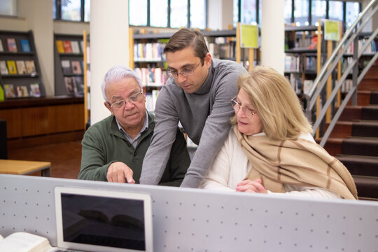 Portrait of aged couple getting knowledges. Serious man and woman sitting in library focused on reading books using computer listening to handsome instructor. Education for mature people concept