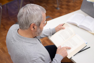 Fototapeta na wymiar Back view of senior man spending time with books. Gray-haired man in glasses reading books in library finding information to study and enjoying active day. Self-education of retired people concept