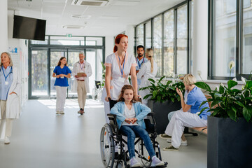 Young nurse pushing little girl on wheelchair at hospital corridor.