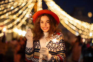 Happy smiling curly woman holding sparklers, posing at winter festive Christmas street fair in European city. Outdoor night portrait. Copy, empty space for text