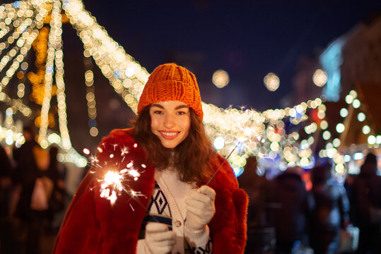 Happy smiling woman wearing knitted beanie hat, faux fur coat, holding sparklers, posing at winter festive Christmas street fair in European city. Outdoor night portrait. Copy, empty space for text
