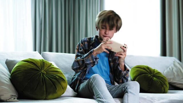 Happy joyful active boy teenager playing mobile phone, video game, having competition, sitting on the sofa holding smartphone. Addicted teenager to video games. Winning, feeling exitement euphoria
