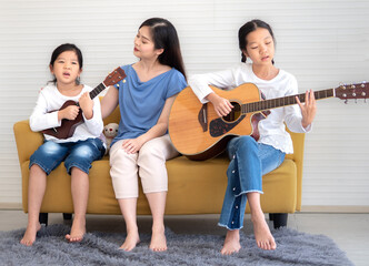 Pretty little girl sitting at couch enjoy playing acoustic guitar with her mother and sibling. Adorable kid playing ukulele with her sister and mom. Asian family guitarist singing together at leisure.