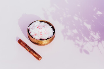 Tibetan singing bowl with floating in water pink peony inside and wooden stick on the pink background. Meditation and Relax. Exotic massage. Minimalism. Direct sunlight and shadows. Selective focus.