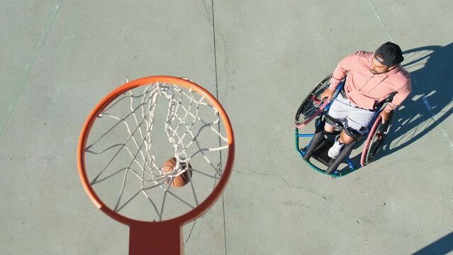 Athletic man in sport wheelchair enjoying streetball game on sunny day at court in city park. Sporty guy with disability making clean throw into hoop and leaving. Amputee sport, achievement concept