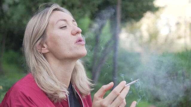 A blonde woman smokes a thin cigarette in a park.