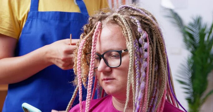Young blonde woman with glasses uses a smartphone, while stylist unwinds her multicolored box braids with special tool, close up. Various female hairstyles.