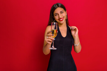 Incredible stylish charming woman with long dark hair and red lips posing with wine over red background. Studio portrait of beautiful woman celebrating New Year, Christmas, birthday