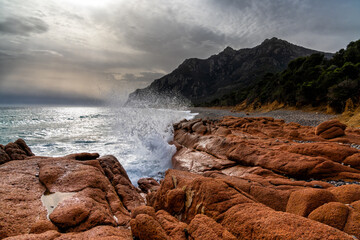 rugged coast in Sardinia with waves crashing onto red granite boulders in the foreground