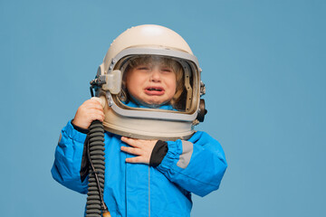 Portrait of little boy, child posing in astronaut costume over blue studio background. Feeling tired, crying