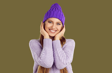 Portrait of beautiful woman in cozy winter clothes. Happy young fashion model in warm purple knitted hat and lavender sweater standing isolated on dark green background, smiling and looking at camera