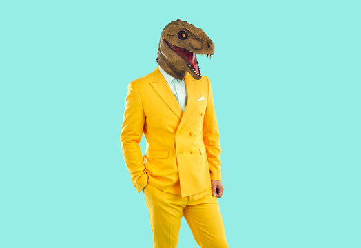 Portrait Of Guy In Funky Costume With Dinosaur Face Posing In Studio. Man In Stylish Bright Yellow Suit And Funny Ugly T Rex Mask Standing With Hand In Pocket On Blue Background. Crazy Fashion Concept