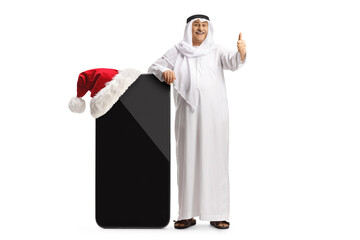 Mature arab man in traditional clothes leaning on a big mobile phone with a santa hat and gesturing...