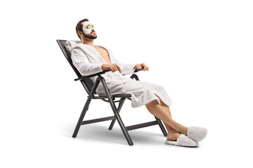 Man in a bathrobe with a face mask and cucumber on eyes sitting on a chair - obrazy, fototapety, plakaty