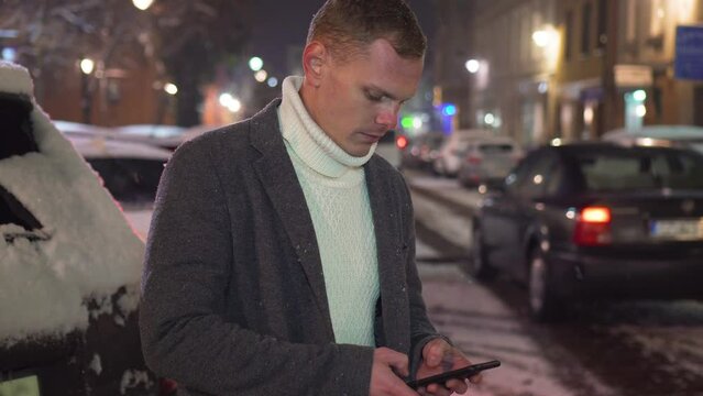 Handsome young man using smartphone on crowded street outdoors under snowfall at night. Checks mail, chats or news on Internet. Posting social media, online shopping, texting, e commerce