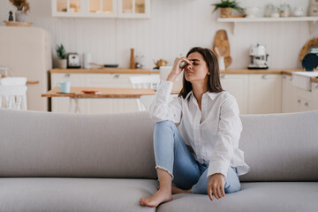 Exhausted brunette hispanic young adult woman in white shirt and blue jeans sitting on sofa eyes closed touches forehead feels fatigue, headache. Tired Caucasian housewife relaxing home. Fatigue - Powered by Adobe