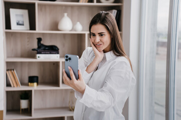 Shocked successful brunette woman at home in white shirt makes video call with wide opened eyes and mouth against blurry interior. Excited pretty girl receives good news, she got a job. Amazed blogger