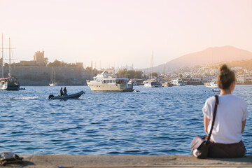 A girl in a white T-shirt sits with her back on the pier against the backdrop of a boat, ship and yachts, mountains and an old fortress