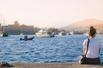 Fototapeta premium A girl in a white T-shirt sits with her back on the pier against the backdrop of a boat, ship and yachts, mountains and an old fortress