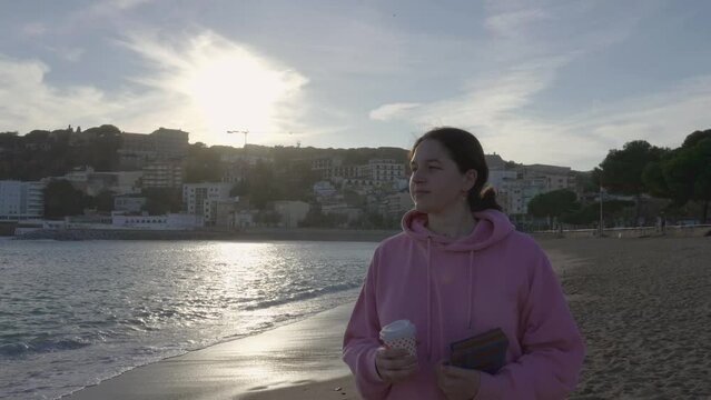 woman walking towards the camera on the beach with a coffee and a book on a winter day wearing a sweater