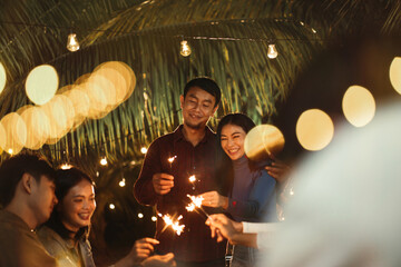 Happy celebrating anniversary party, Asian couple with group friends and having fun with firework sparklers at night party outdoor. Friendship and celebration concept.