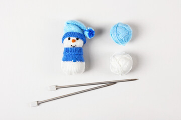 Knitted snowman in a blue hat and sweater with two balls of thread and knitting needles on a gray background. Knitted Christmas decorations