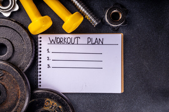 Fitness or bodybuilding background. Old hard iron dumbbells, small colorful dumbbells,  notepad with workout plan list. Back to gym concept, weight lifting sport flat lay top view copy space  
