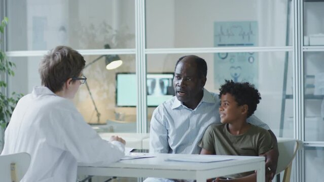 Little African American boy sitting with father in pediatric clinic and listening to female doctor while getting medical consultation