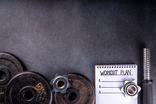 Fitness or bodybuilding background. Old hard iron dumbbells, small colorful dumbbells,  notepad with workout plan list. Back to gym concept, weight lifting sport flat lay top view copy space  
