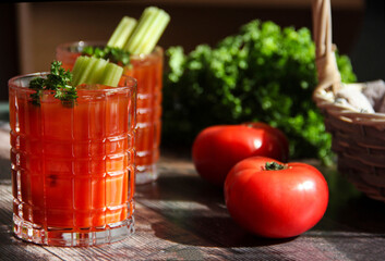 Bloody Mary cocktail with ice cubes, celery sticks and parsley leaves. Tomato juice. Red tomato cocktail in transparent glasses on a wooden table on a sunny day with tomatoes on the table. Close up.