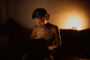lonely man in a dark room at night with a naked torso sits with a laptop on the couch.