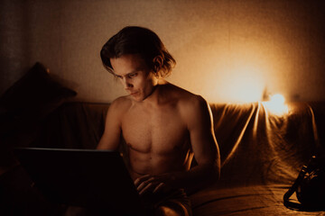 lonely man in a dark room at night with a naked torso sits with a laptop on the couch.
