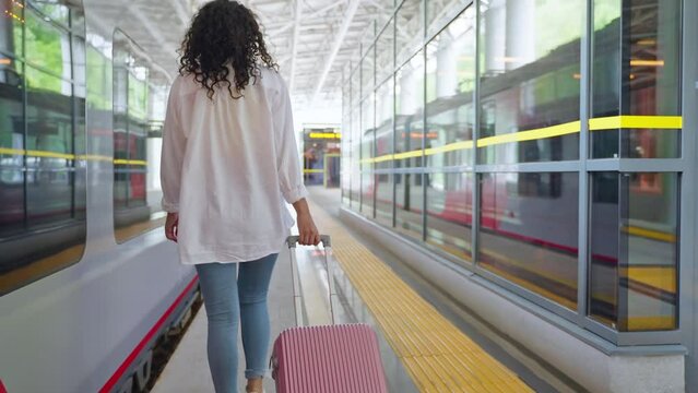 woman traveller is walking on train station, rear view, tourist is rolling suitcase
