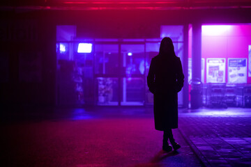dark silhouette of a girl dressed in a long coat against the background of a night city with advertising showcases - obrazy, fototapety, plakaty